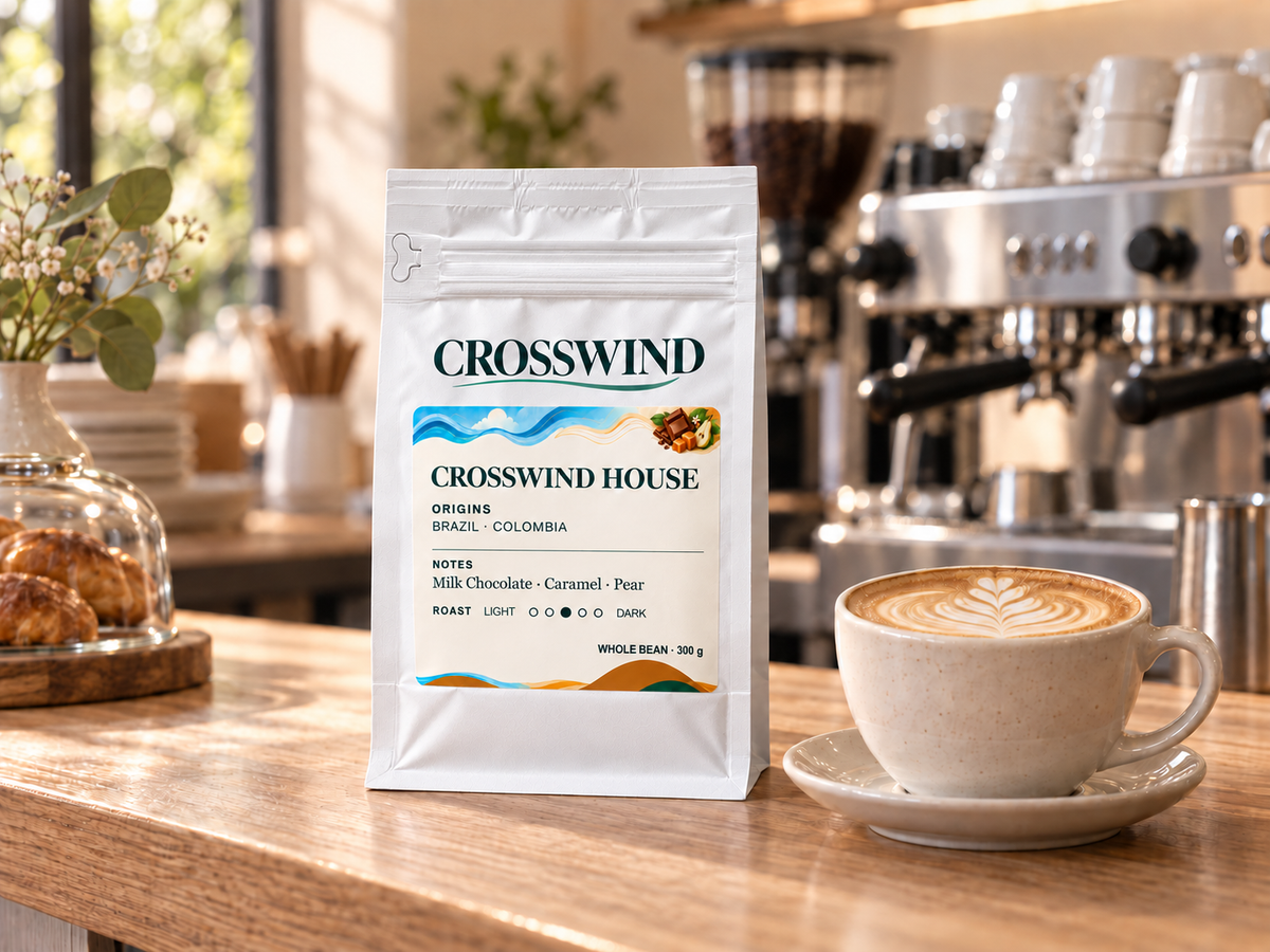 Illustrative sealed Crosswind House 300 g retail bag beside a cappuccino and espresso machine in a cafe